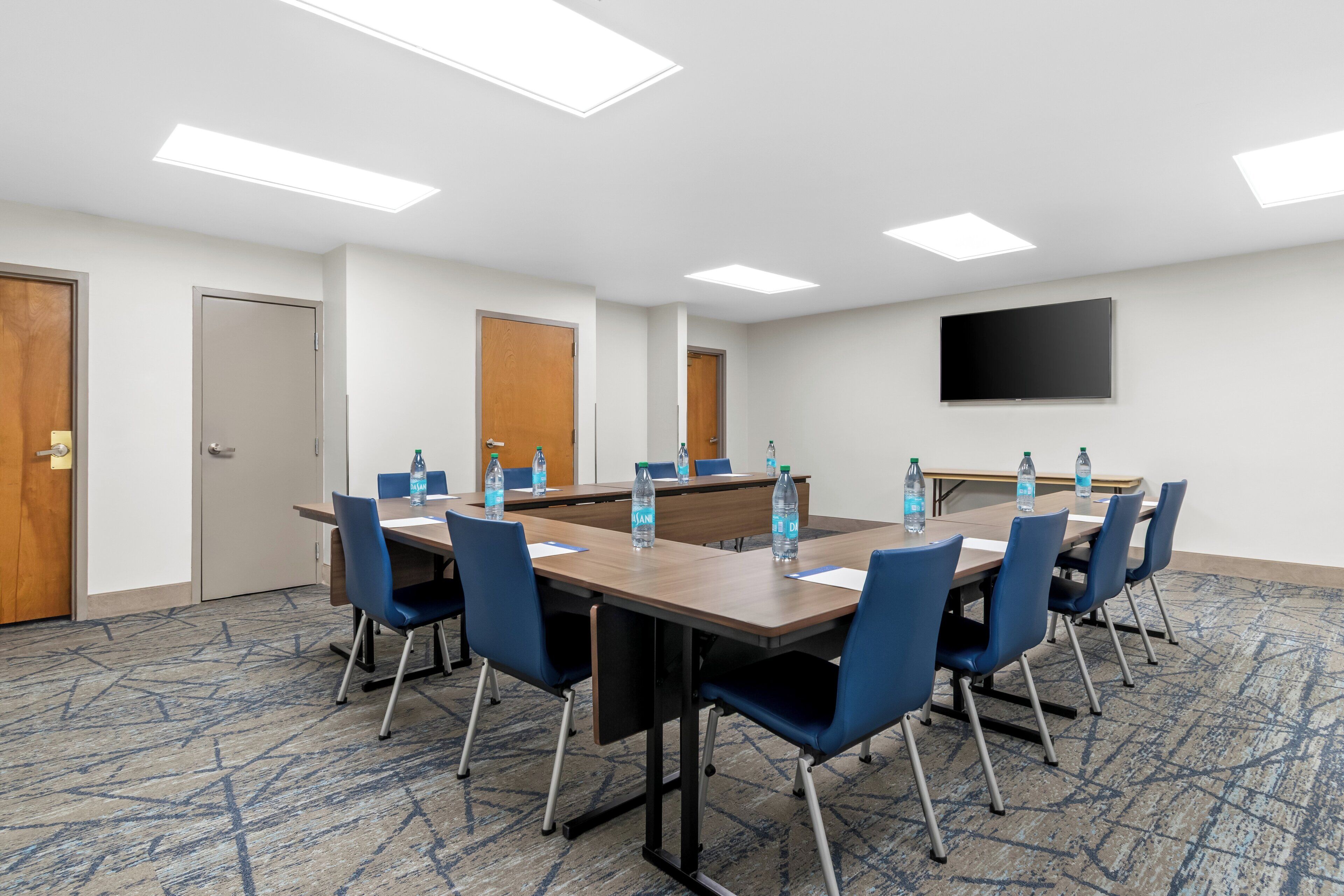 A conference room with a long table, blue chairs, water bottles, and a flat-screen TV on the wall.