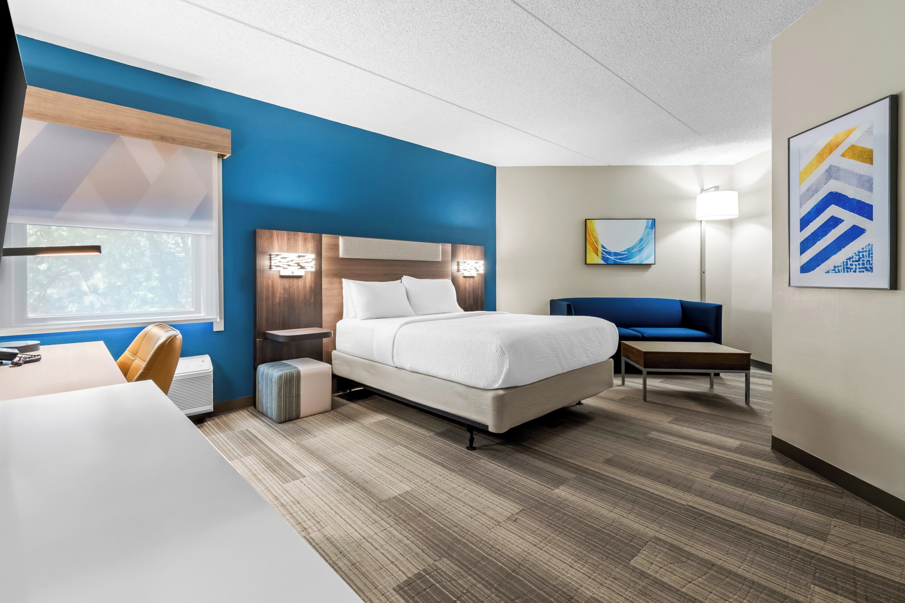 A hotel room with a bed, a desk, a chair, a sofa, a lamp, and artwork on the walls.