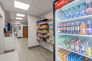 En lys og indbydende gang med et Coca-Cola køleskab fyldt med drikkevarer, hylder med snacks og en trædør for enden.