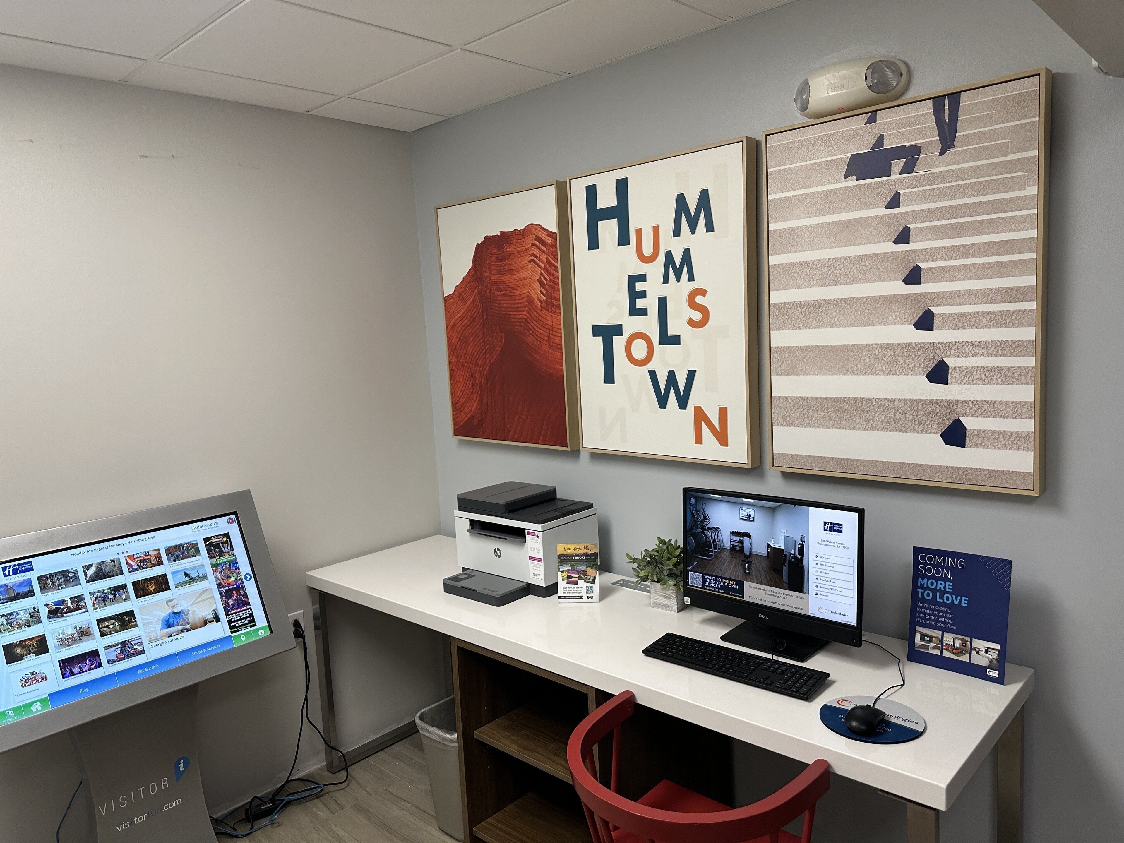 A modern office desk with a computer, printer, and keyboard. There are two framed posters on the wall, one with the text "HUMES TOWN" and another with a geometric design. A monitor is on the desk, and a trash can is visible.