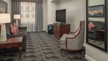 Una habitación de hotel con un sofá, una silla, un televisor, un aparador y un espejo grande.
