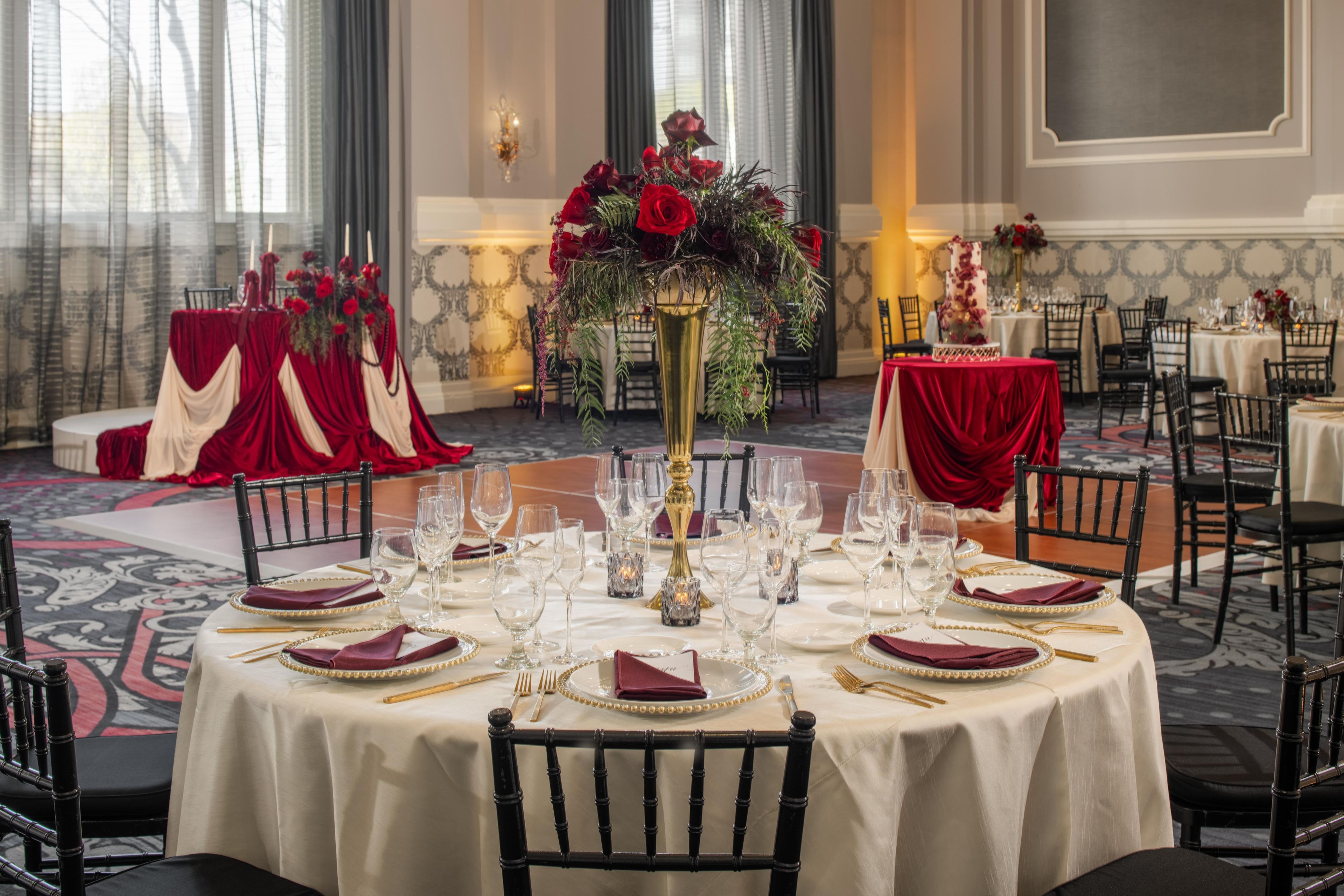 A formal dining room with round tables, red drapery, and a centerpiece of red roses.