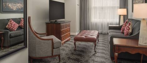 A modern hotel room with a large flat-screen TV, a grey sofa, a wooden cabinet, and a patterned carpet.