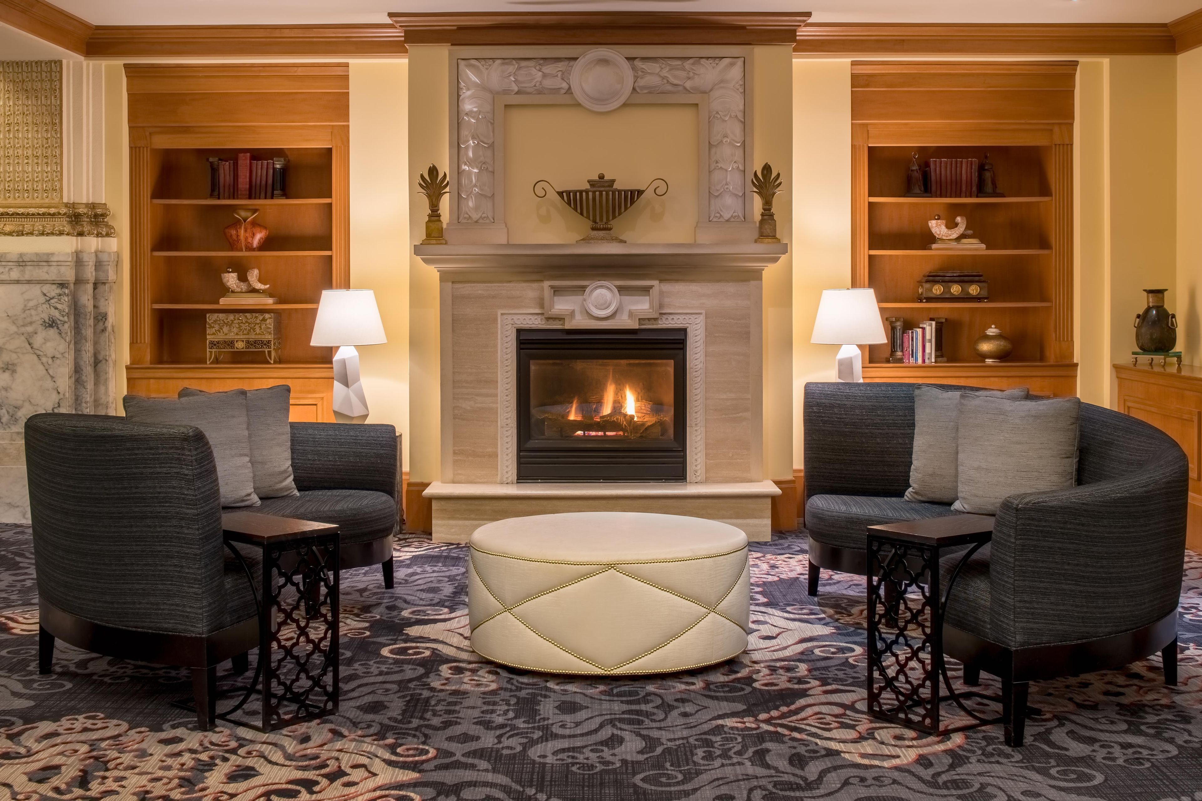 A room with a fireplace, two armchairs, a round ottoman, and bookshelves with decorative items.