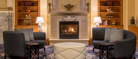 A room with a fireplace, two armchairs, a round ottoman, and bookshelves with decorative items.
