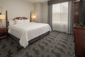 A hotel room with a large bed, a wooden headboard, two bedside lamps, a flat-screen TV, and a large window with sheer curtains. - Embassy Suites by Hilton Portland Downtown (Portland)