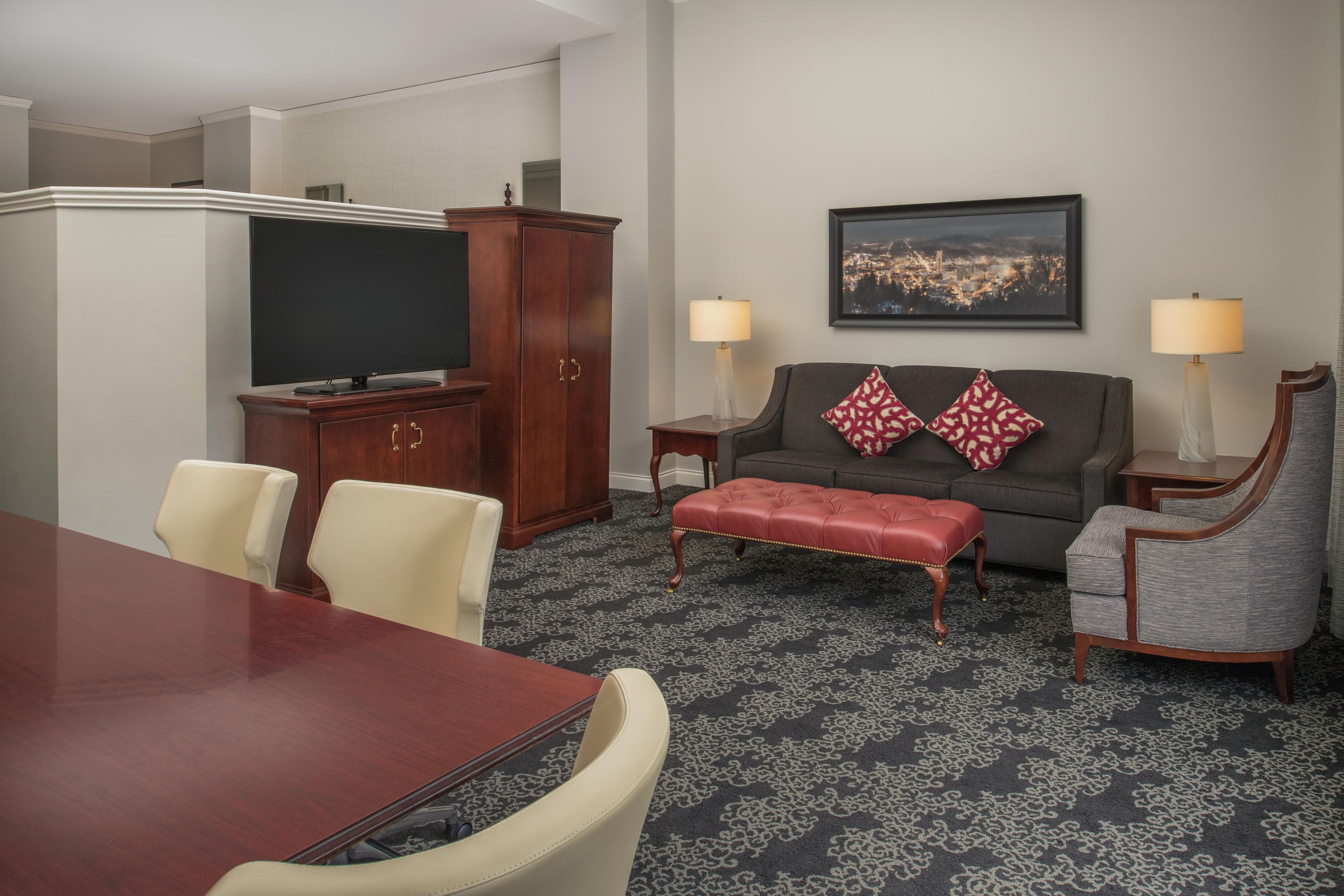 Quarto de hotel com mesa de jantar, cadeiras, sofá, um banquinho vermelho, televisão, armário e duas luminárias.