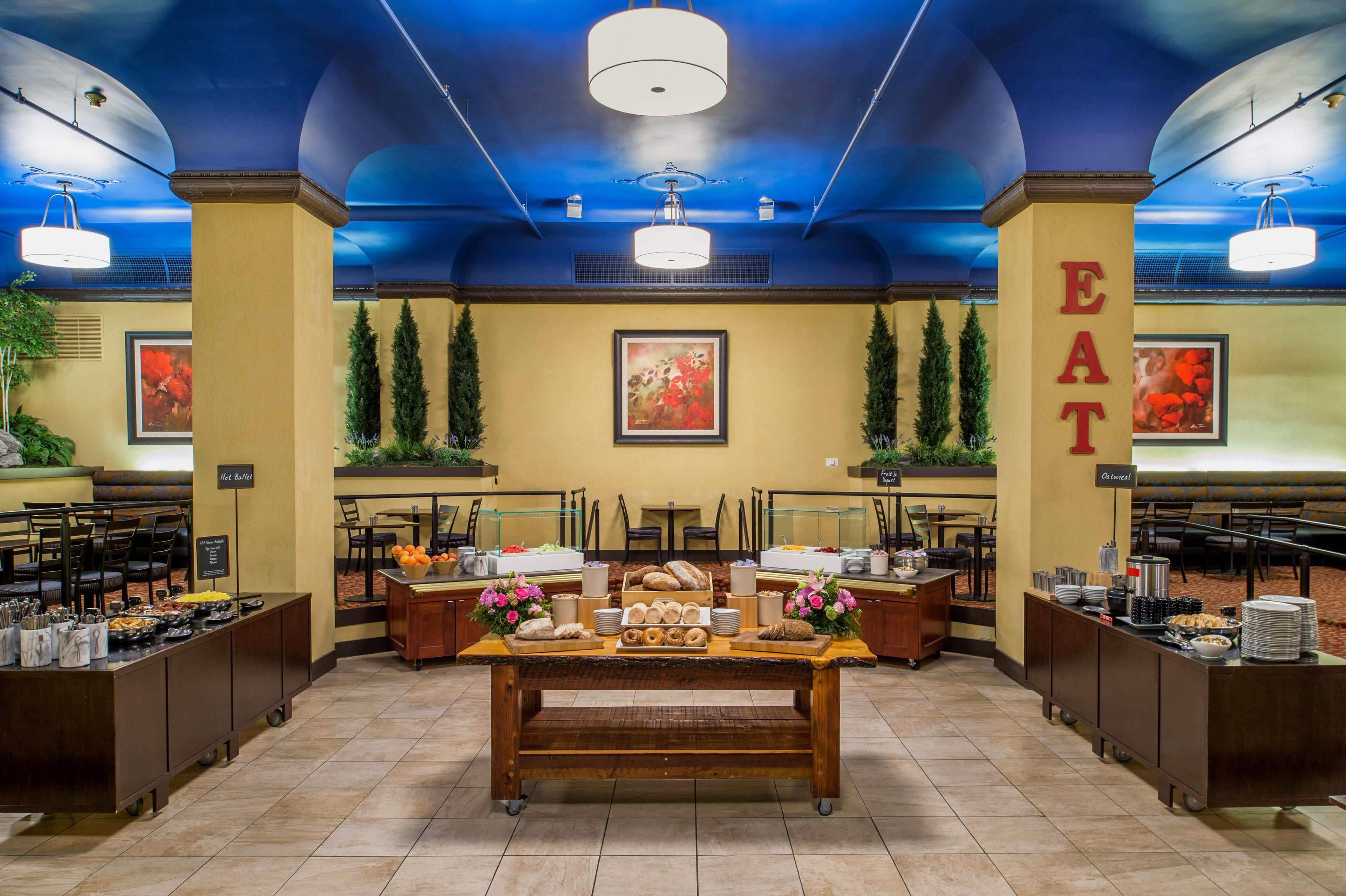 A hotel breakfast area with a buffet setup, seating arrangements, and a prominent 'EAT' sign on the wall.
