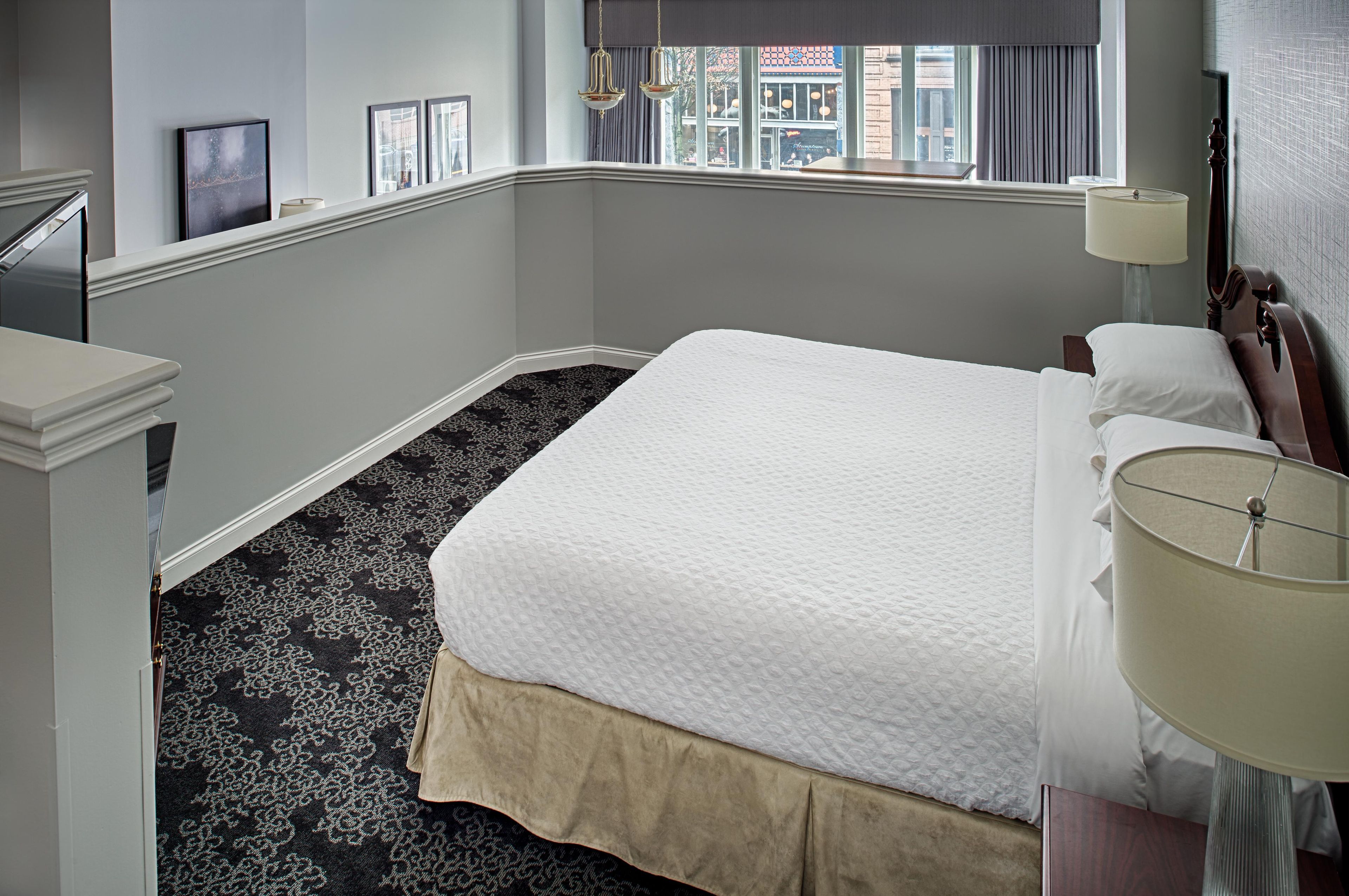 A hotel room with a bed, a nightstand, a lamp, a window with a view of the street, and a patterned carpet.