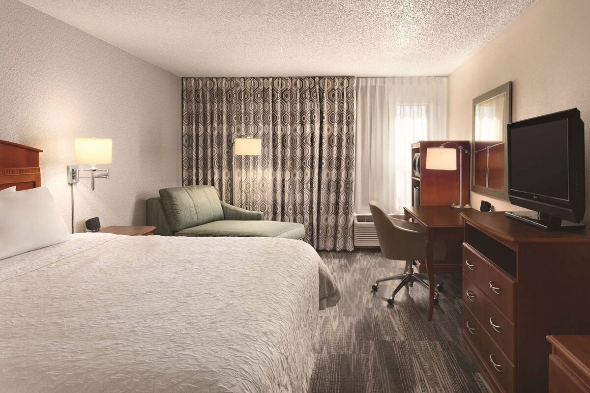 A hotel room with a large bed, a desk with a chair, a television, and a window with curtains.