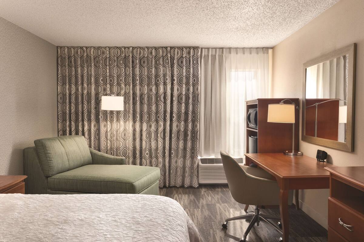 A hotel room with a bed, a desk, a chair, a dresser, a mirror, a lamp, and patterned curtains.