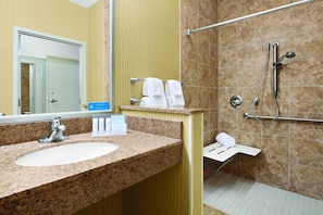 A bathroom with a granite countertop sink, a mirror, a towel rack with two white towels, a shower with a handheld showerhead, and a grab bar.