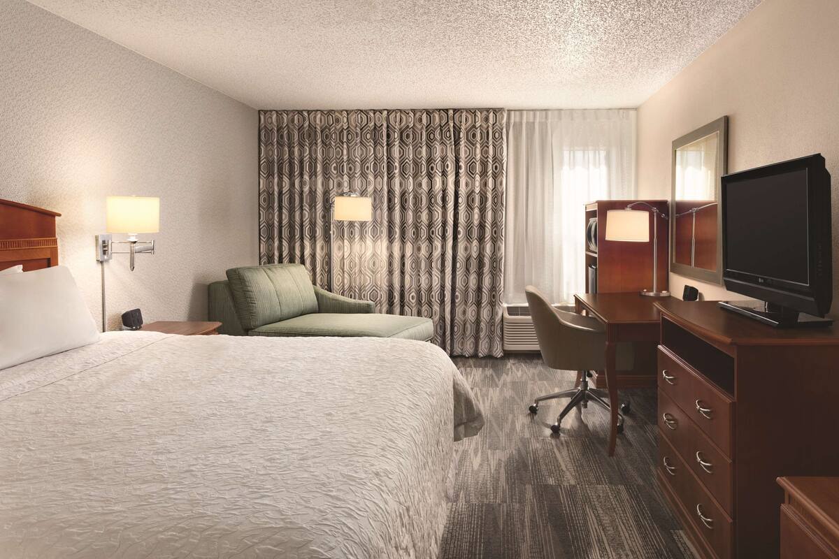 A hotel room with a large bed, a desk, a chair, a television, and a wardrobe.