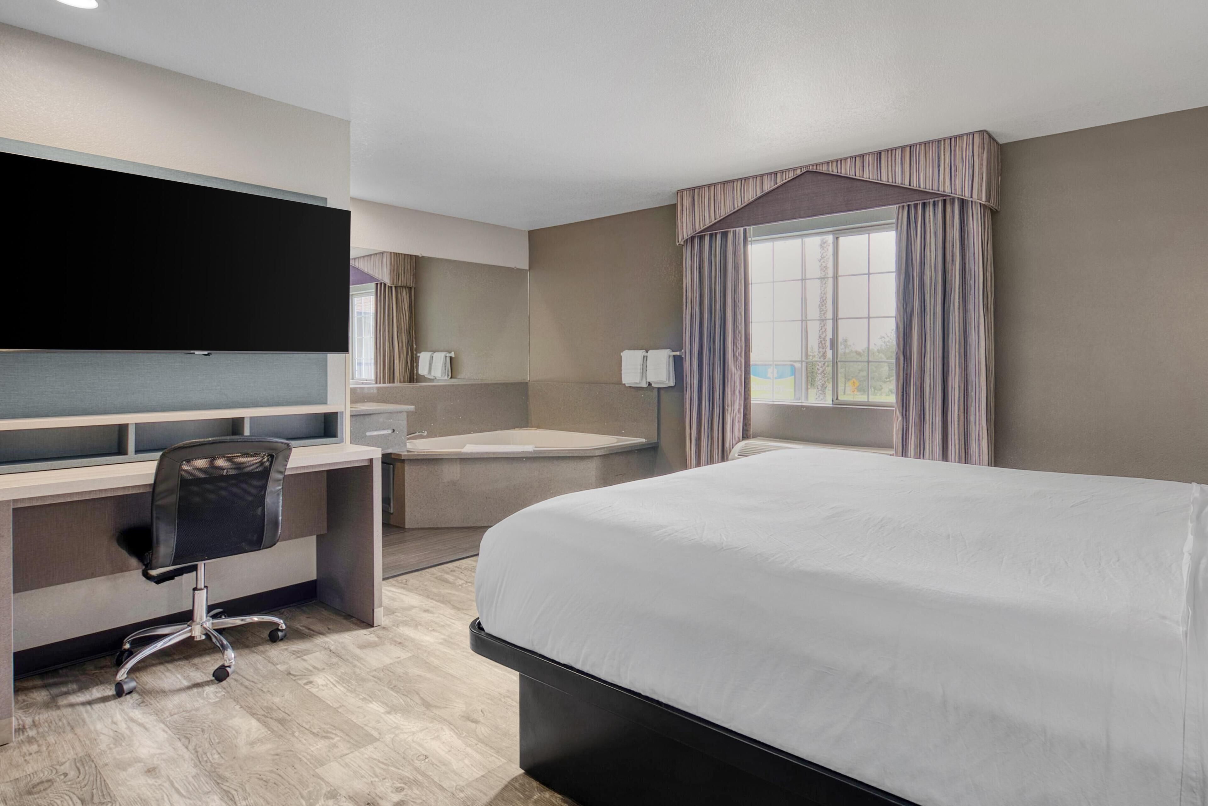 Habitación de hotel con una cama grande, un escritorio con silla, televisor de pantalla plana, bañera y ventana con cortinas.
