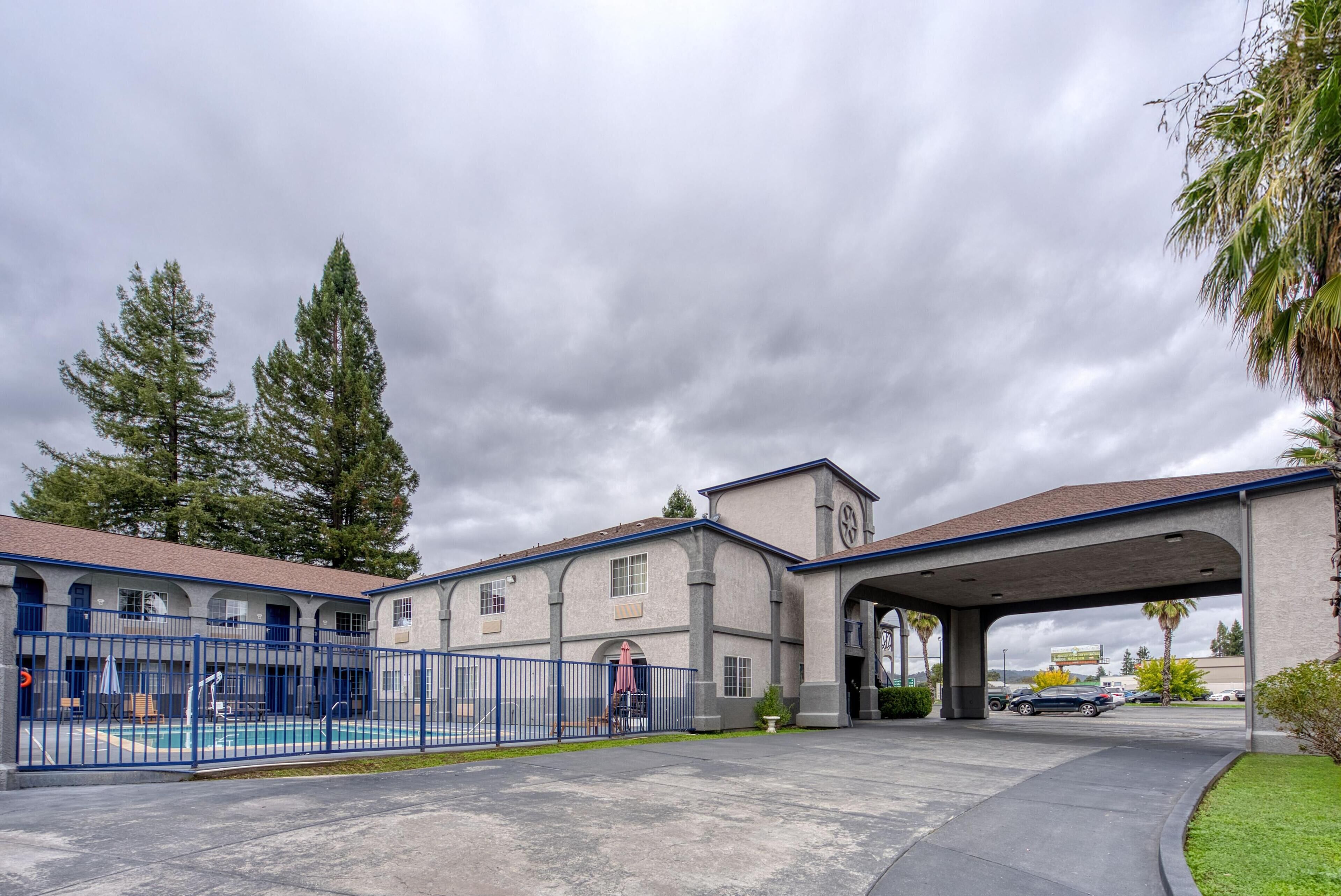A motel with a swimming pool, covered parking, and a large building with a clock tower.