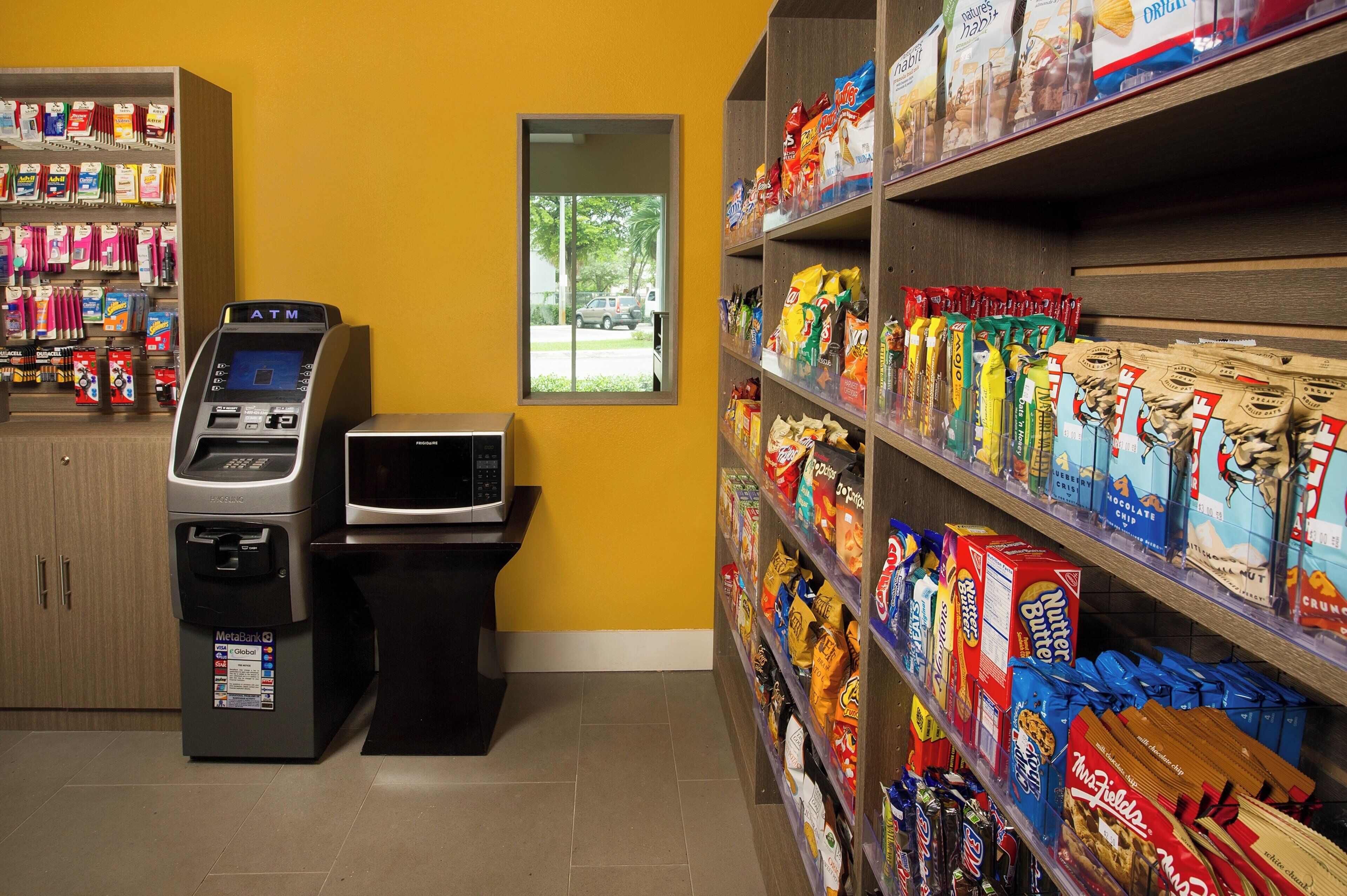 A self-service kiosk area with a payment terminal, a microwave, and shelves stocked with snacks and packaged goods.