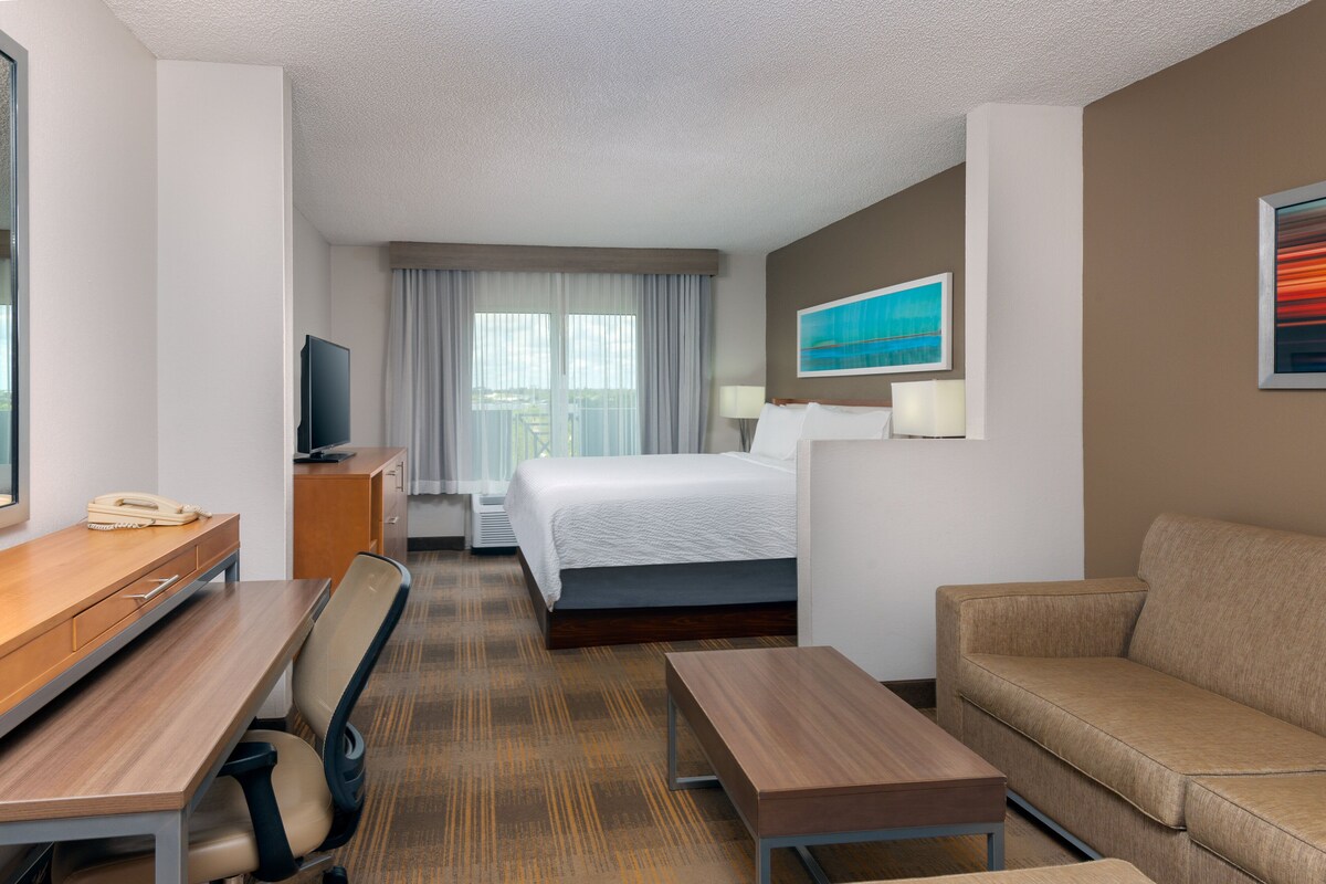 A hotel room with a bed, a desk with a telephone, a chair, a sofa, a television, and a window with curtains.