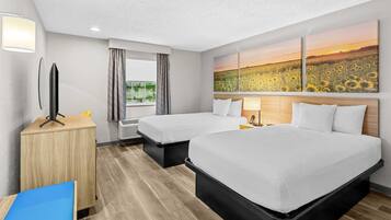 Habitación de hotel con dos camas, un escritorio, una ventana con cortinas, un radiador y una pared con un cuadro de un girasol.
