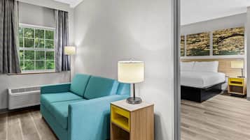 A hotel room with a sofa, a bedside table with a lamp, a bed, and a window with curtains.