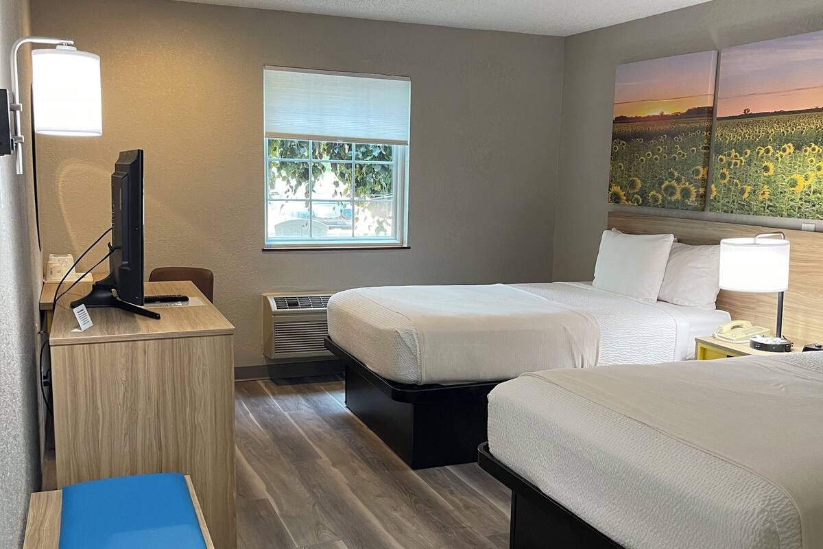 A hotel room with two beds, a desk with a computer, a window with blinds, and a painting of a sunset over a field.