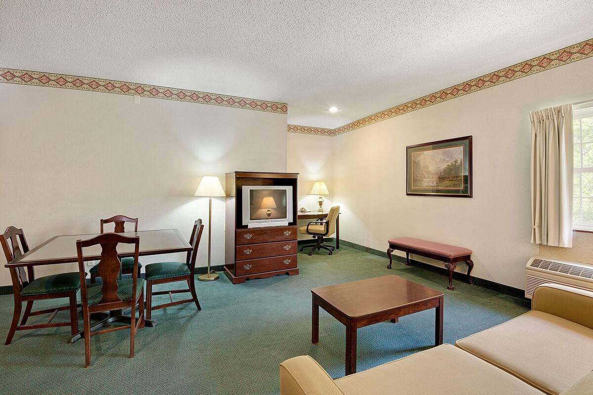 A hotel room with a dining table, chairs, a television, a desk, a computer, a chair, a sofa, and a window with curtains.