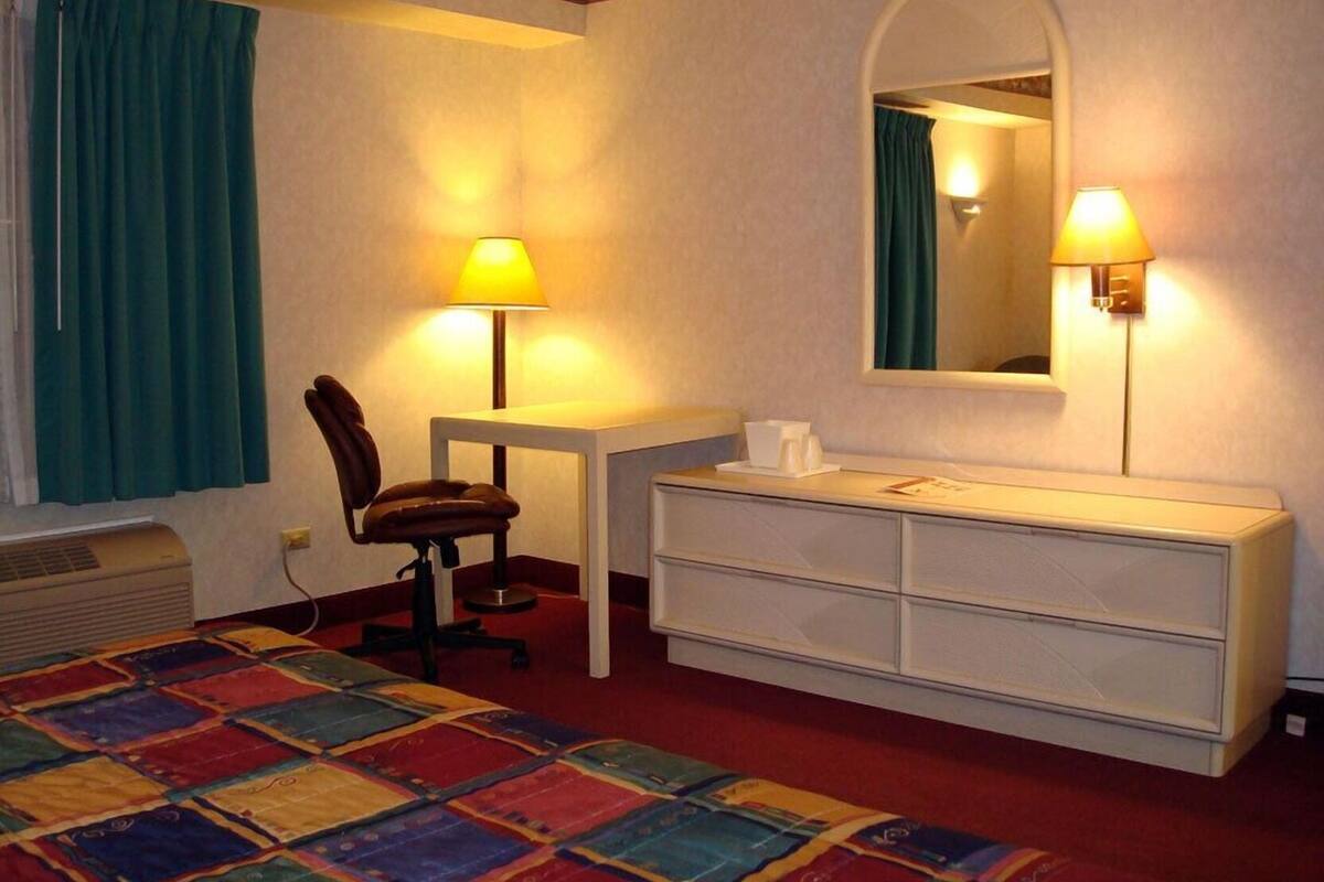 A hotel room with a bed, a desk, a chair, a dresser, two lamps, and a window with curtains.