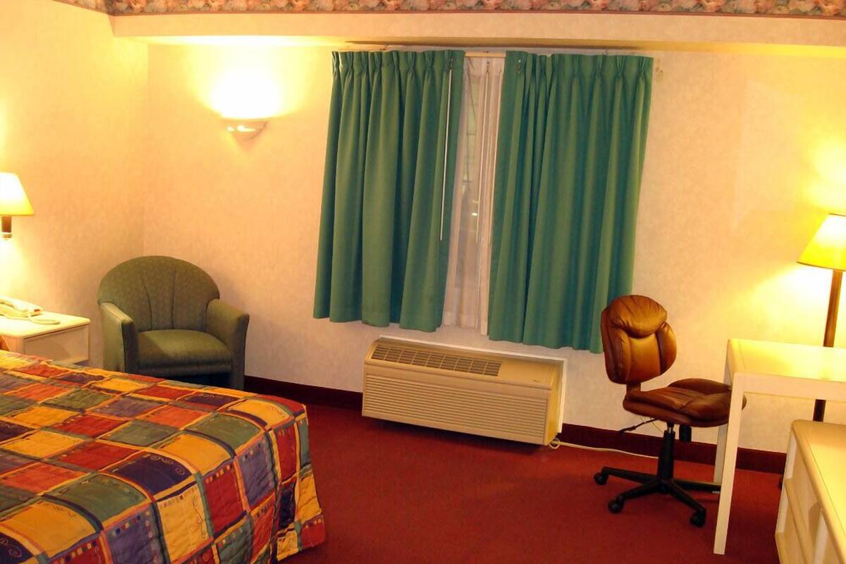 A hotel room with a bed, a desk, two chairs, a heater, and a window with curtains.