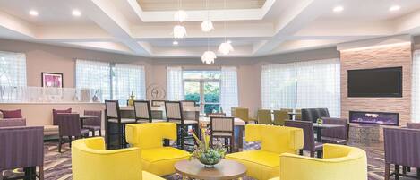 A hotel lobby with yellow armchairs, a round table with a vase, and a flat-screen TV mounted on the wall.