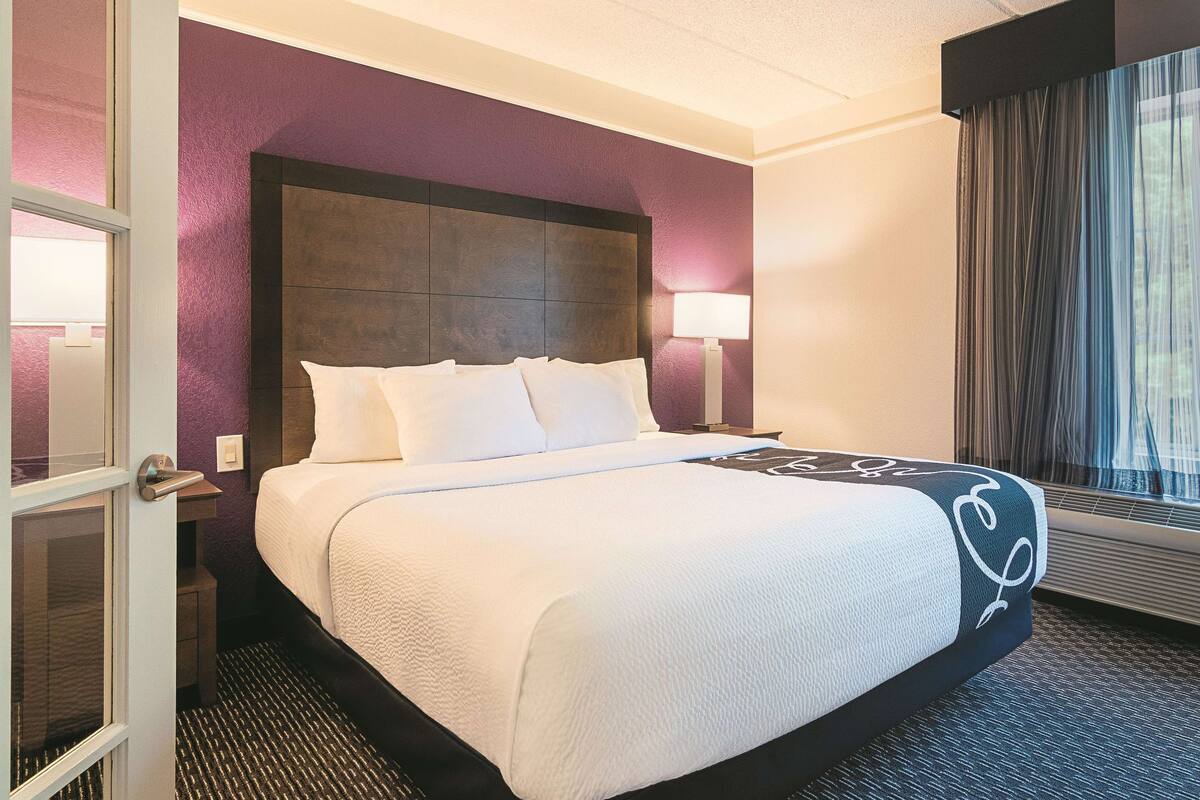 Une chambre d’hôtel avec un grand lit, une tête de lit, une table de chevet avec une lampe et une fenêtre avec des rideaux.