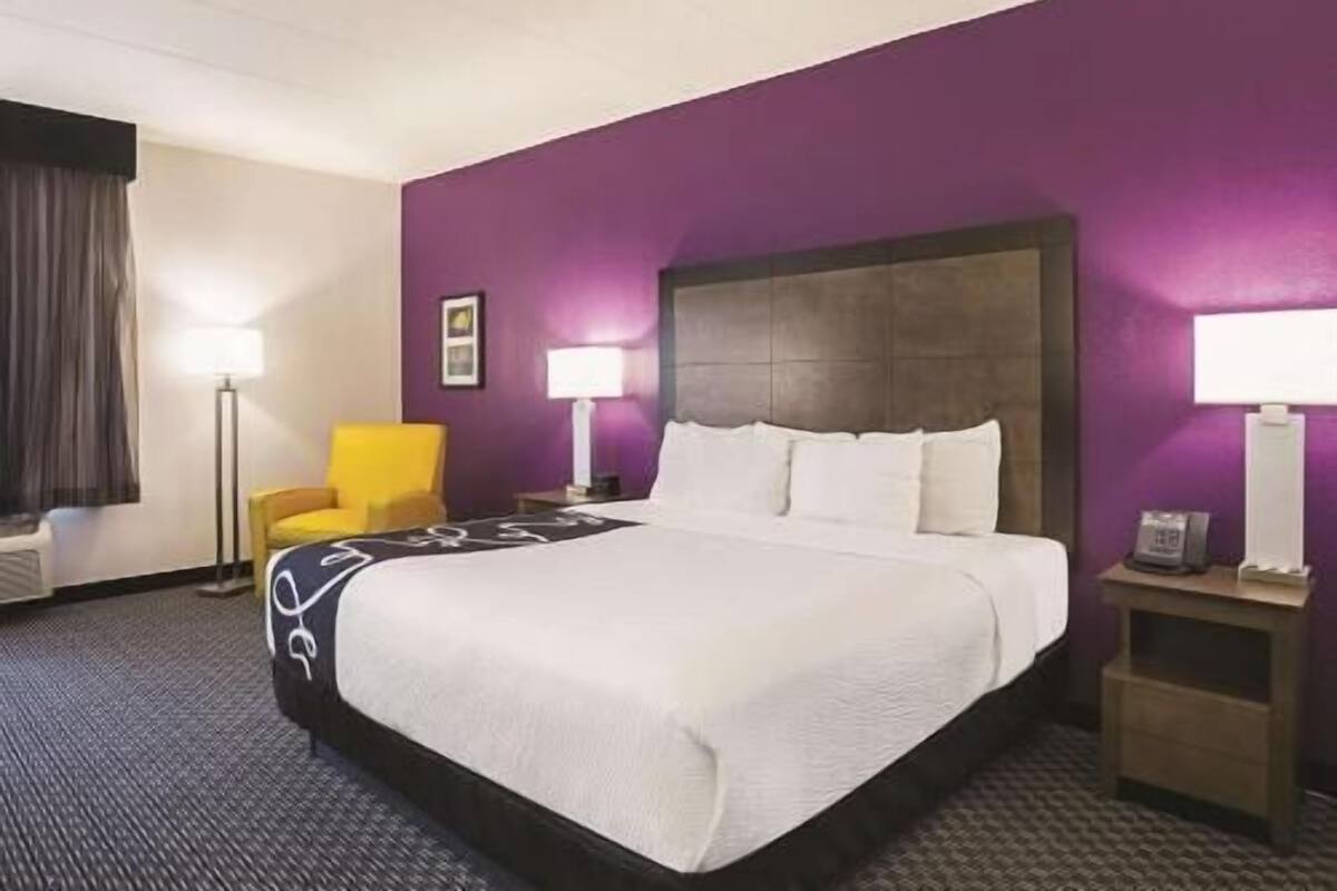 Une chambre d’hôtel avec un grand lit, deux tables de chevet, une chaise et un mur de couleur violette.
