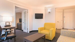 Une chambre d’hôtel équipée d’un bureau, d’un fauteuil jaune, d’un canapé à motifs, d’un téléviseur à écran plat et d’une porte donnant sur une autre pièce.