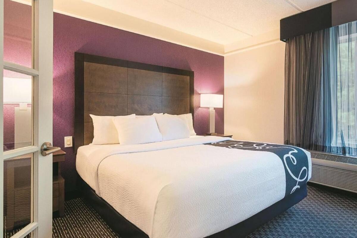 Une chambre d’hôtel avec un grand lit, une tête de lit, une table de chevet, une lampe et une fenêtre avec des rideaux.