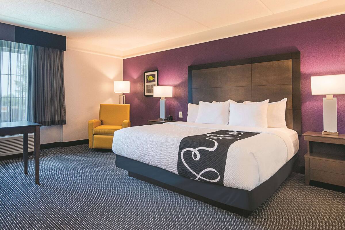 Une chambre d’hôtel avec un grand lit, une chaise, deux tables de chevet avec des lampes et une fenêtre avec des rideaux.