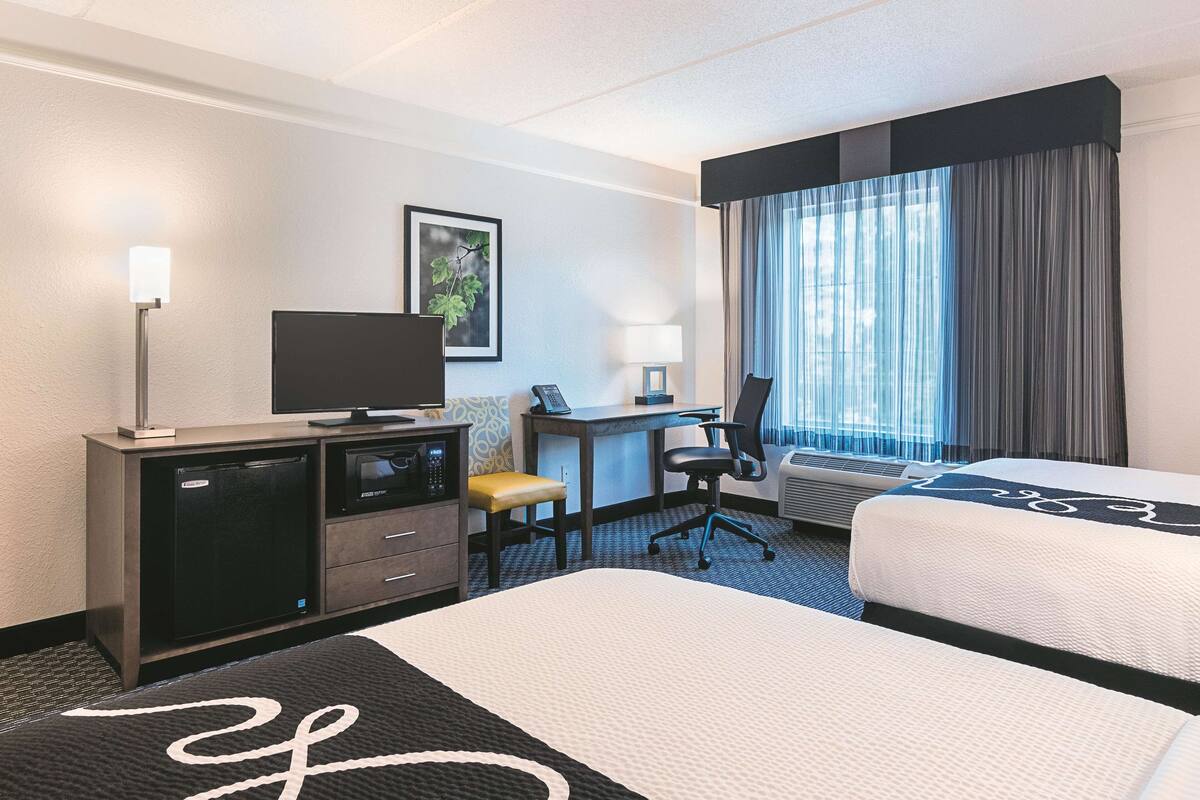 Une chambre d’hôtel avec deux lits, un bureau avec un ordinateur, une télévision, une lampe et une fenêtre avec des rideaux.