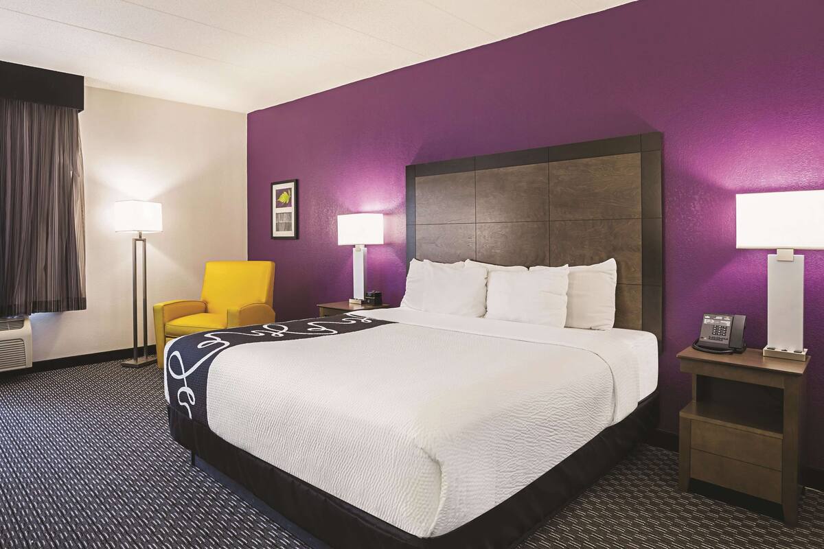Une chambre d’hôtel avec un grand lit, un fauteuil jaune, deux tables de chevet avec des lampes et un mur violet.