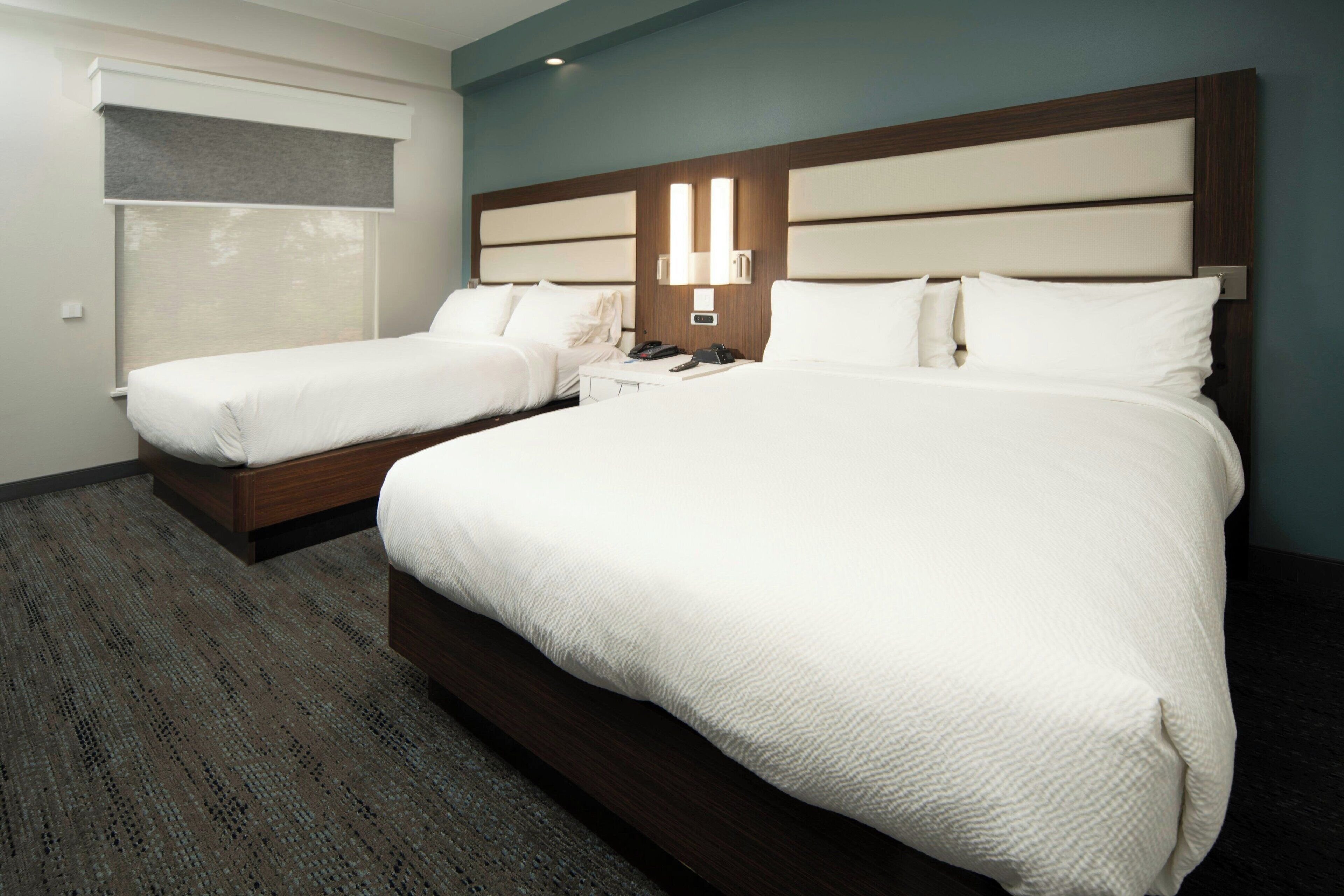 A hotel room with two beds, a nightstand, a lamp, and a window with blinds.