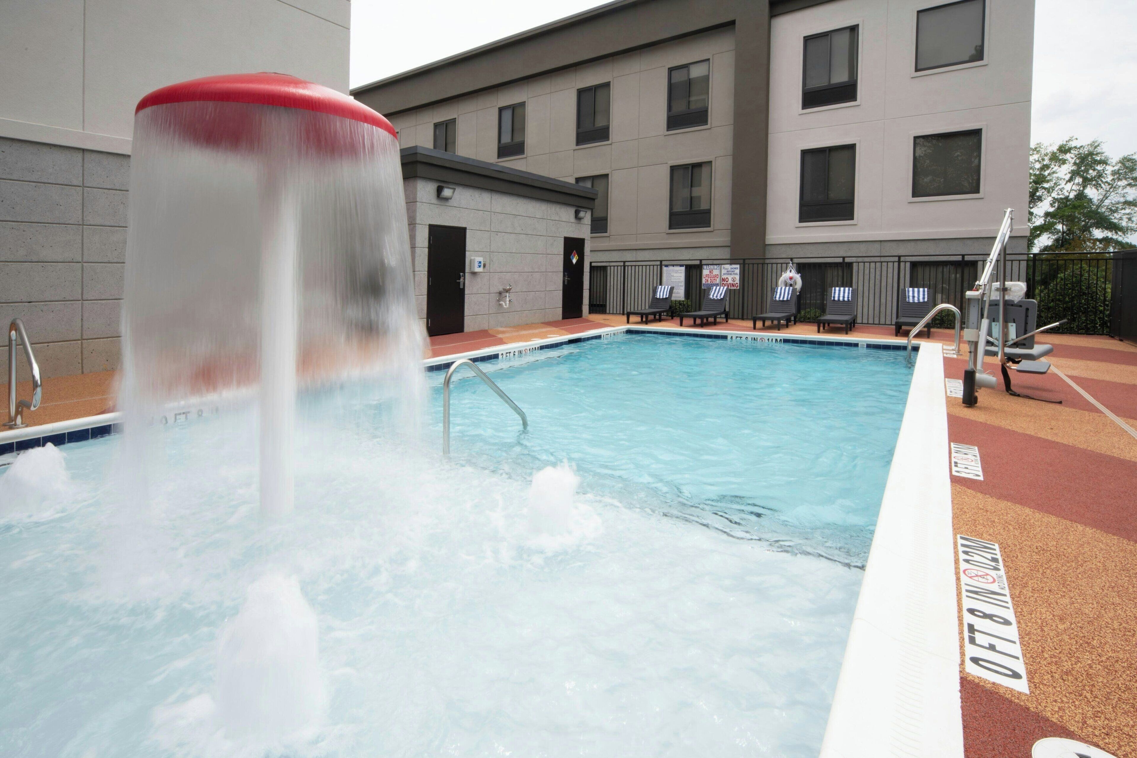 A swimming pool with a water feature, adjacent to a building with multiple windows and a covered area with lounge chairs.