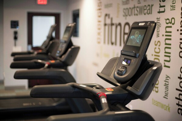 A row of stationary bikes with digital screens, a wall with motivational words, and a door in the background. - Four Points by Sheraton Fayetteville Fort Bragg (Fayetteville)
