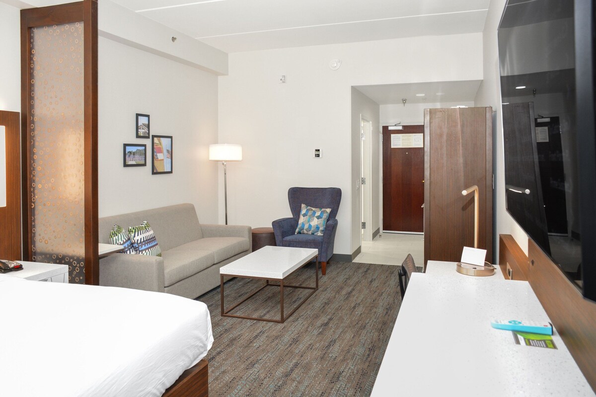 A hotel room with a bed, a sofa, a small table, a chair, and a TV.
