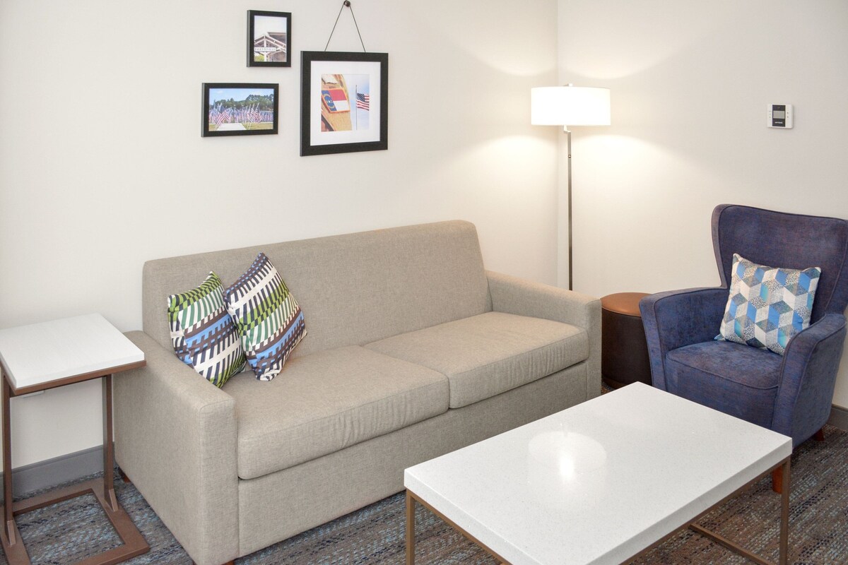 A living room with a beige sofa, a blue armchair, a white coffee table, and a floor lamp. There are framed pictures on the wall.