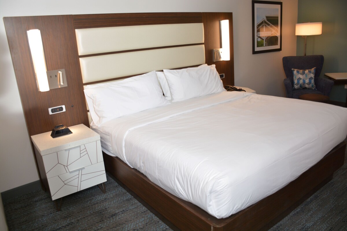 A large bed with a wooden headboard, a bedside table with a geometric design, a lamp, and a chair with a patterned cushion.