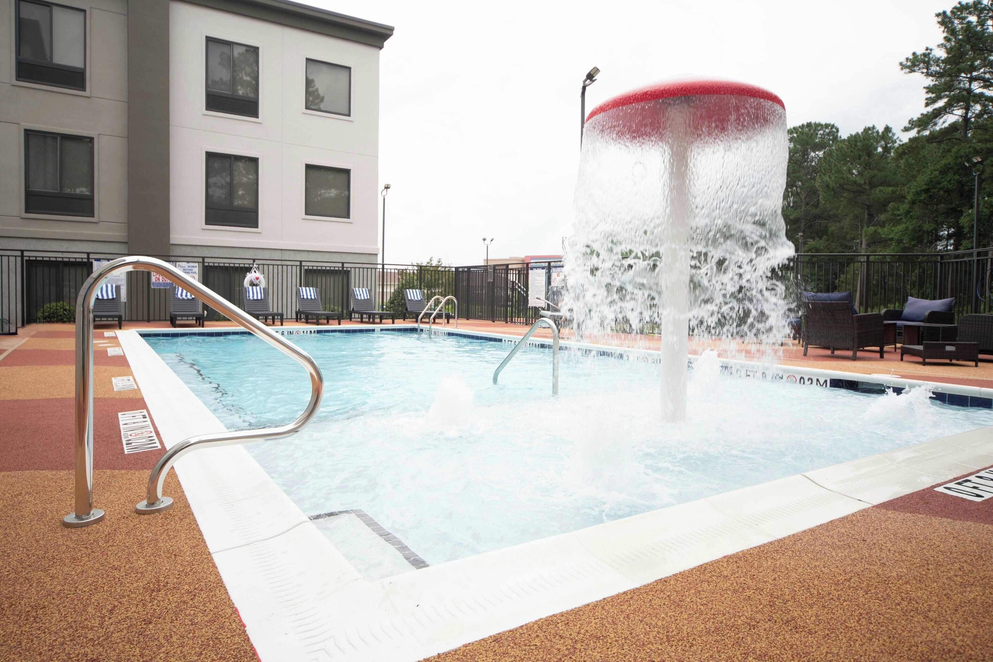 A swimming pool with a water fountain, lounge chairs, and a building in the background.