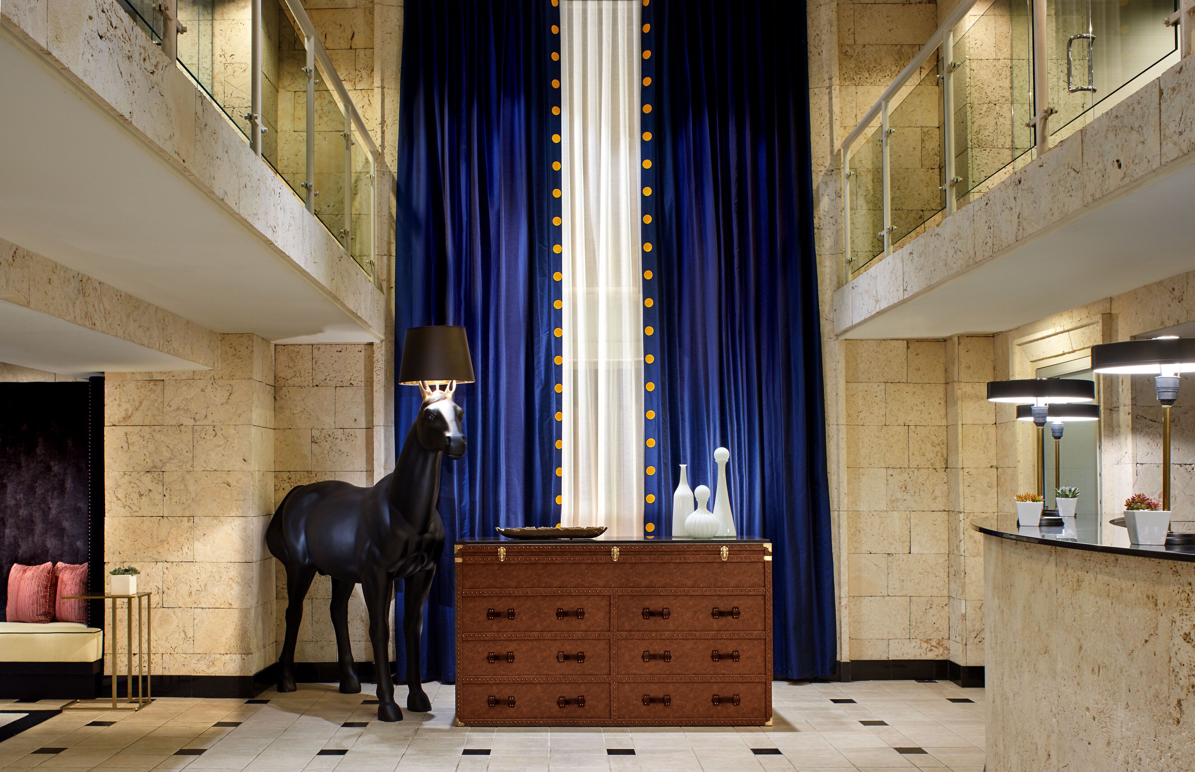 Un hall d’entrée luxueux avec un sol en marbre, un grand rideau bleu, une commode en bois et une statue de cheval noir.