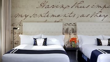 Habitación de hotel con dos camas, cada una con ropa blanca y detalles azul marino. Se observa una pared con un diseño decorativo de escritura y una mesita con un jarrón de flores.