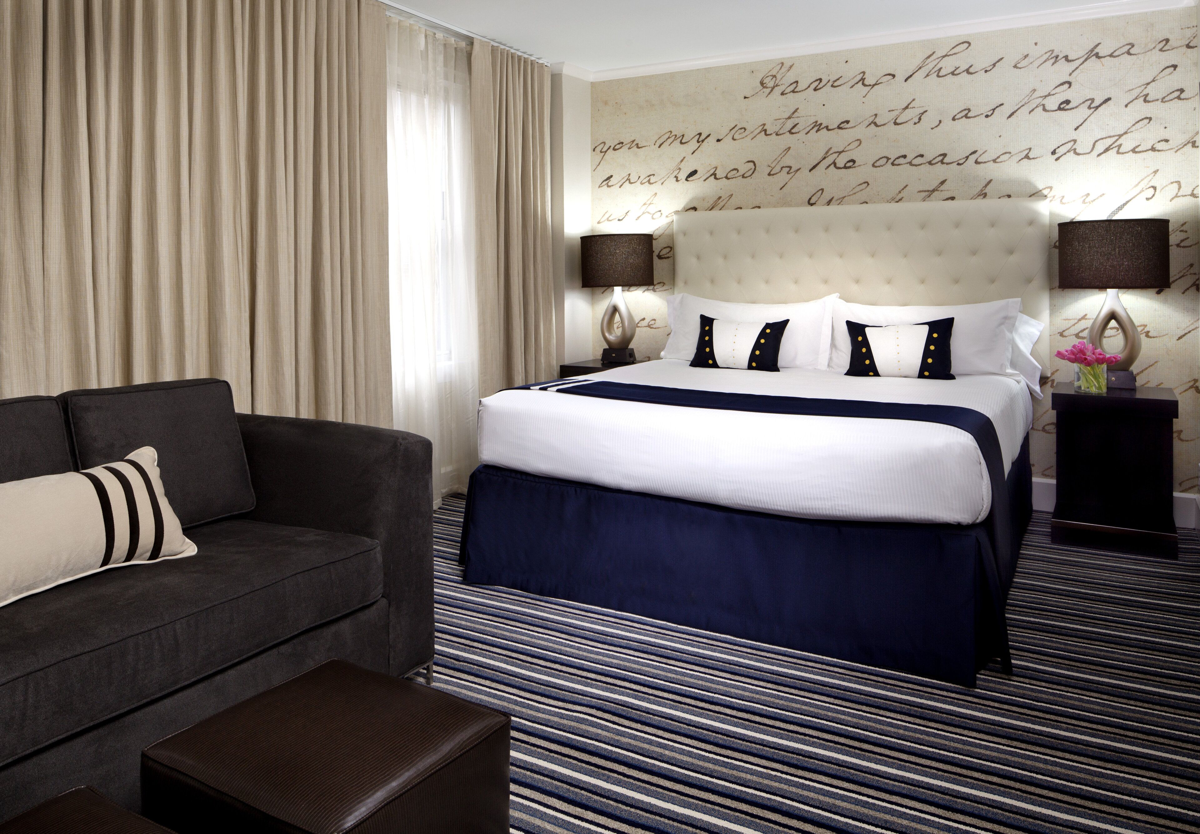 A hotel room with a large bed, a dark sofa, a striped carpet, and a wall with decorative script.