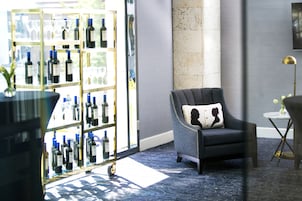 A room with a dark grey armchair, a glass display case with wine bottles, a marble column, and a small round table with a lamp.