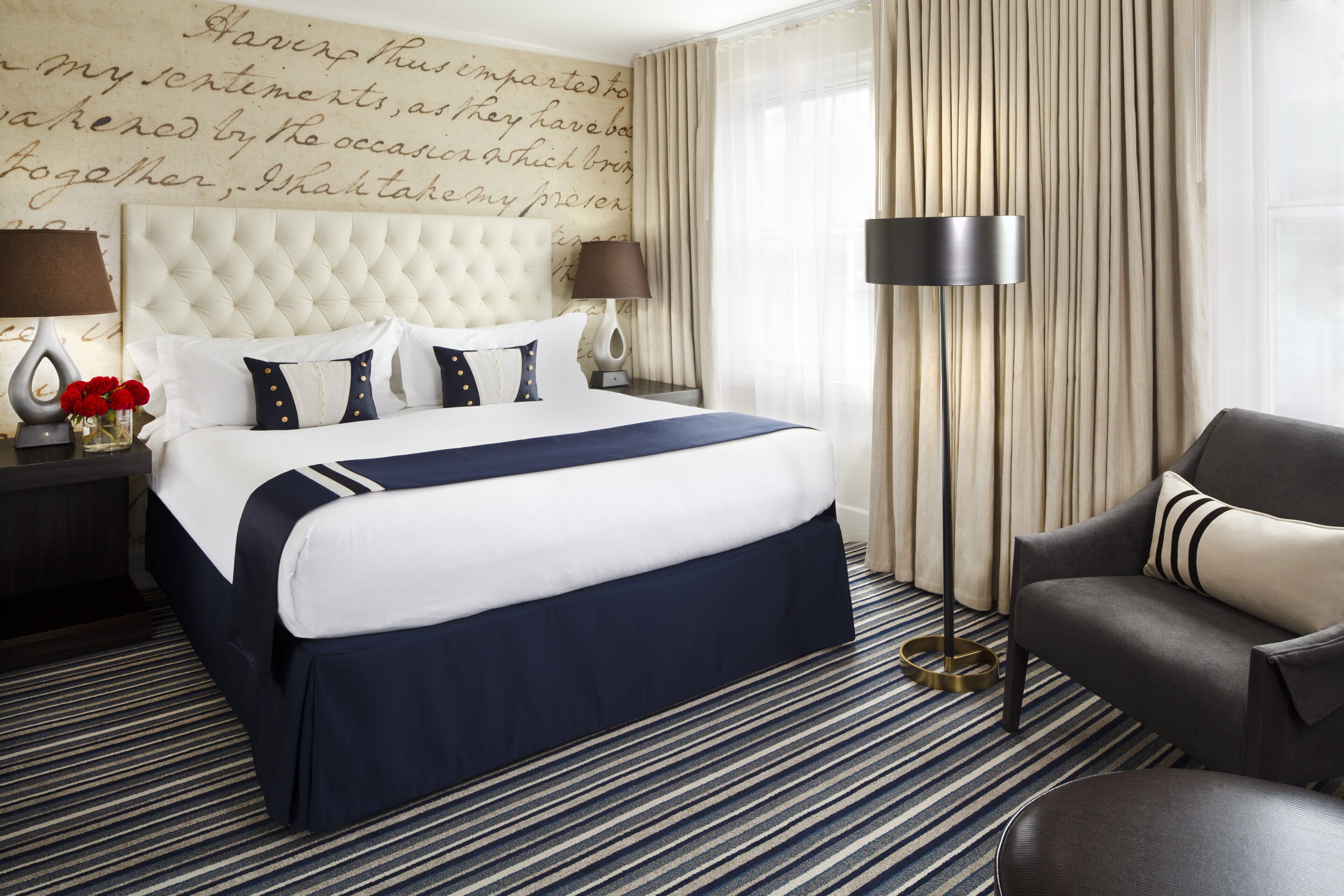 Une chambre d’hôtel avec un grand lit, deux tables de chevet, une chaise, une lampe sur pied et un mur décoratif orné d’inscriptions.