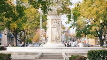A public square with a central monument featuring inscriptions, surrounded by trees and benches.