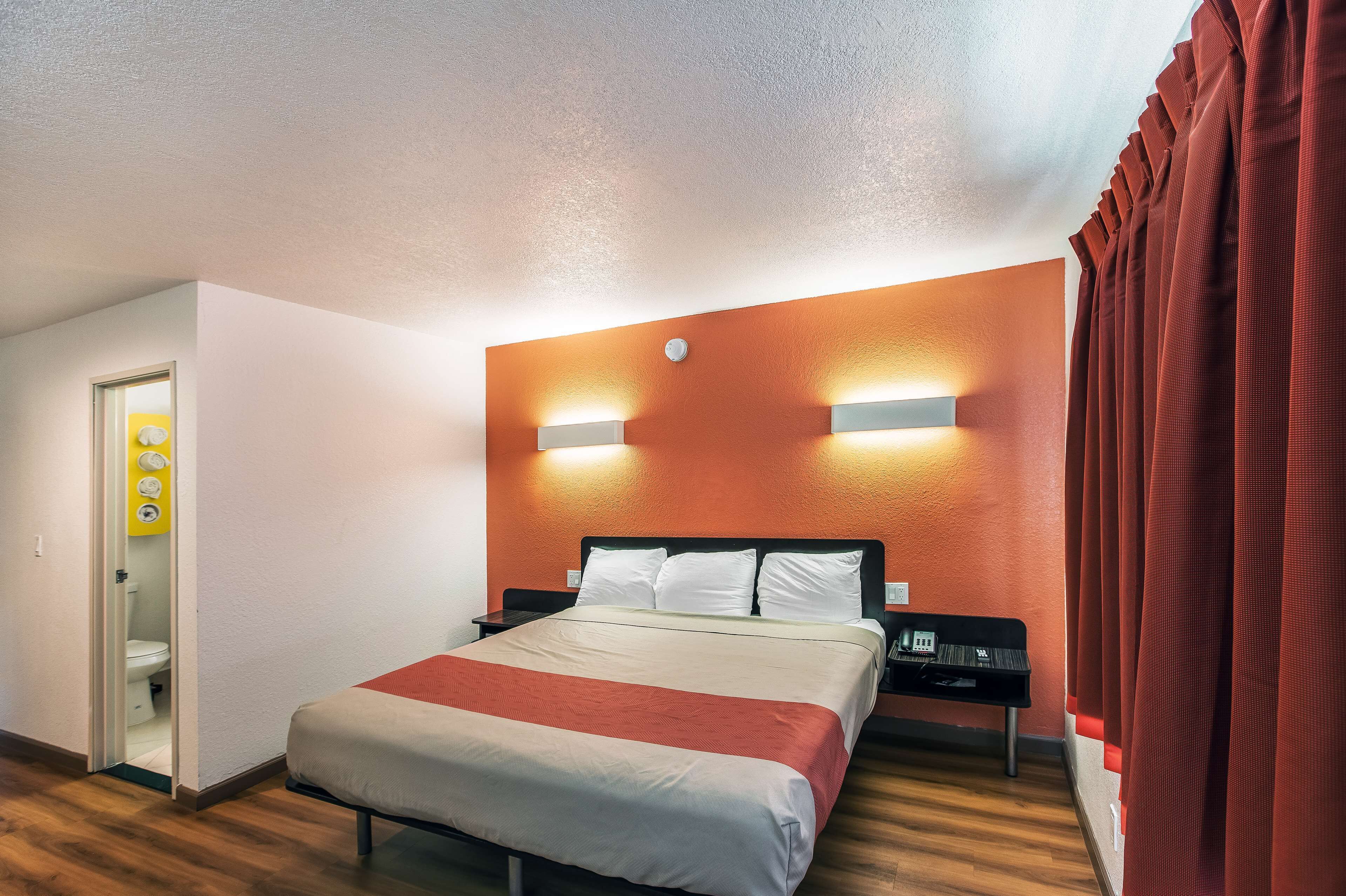 A hotel room with a bed, a nightstand, a wall-mounted light, and a door leading to a bathroom.