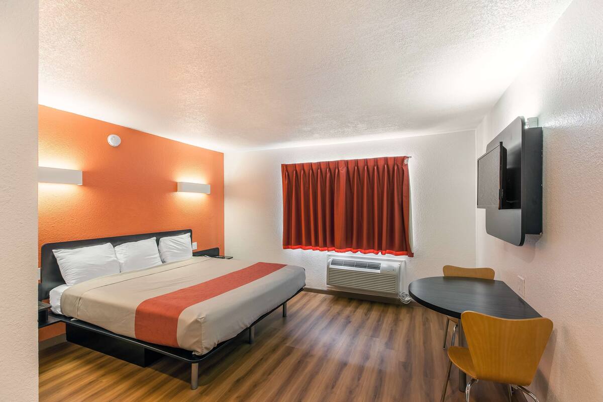 A hotel room with a bed, a small table with two chairs, a TV mounted on the wall, and a window with red curtains.